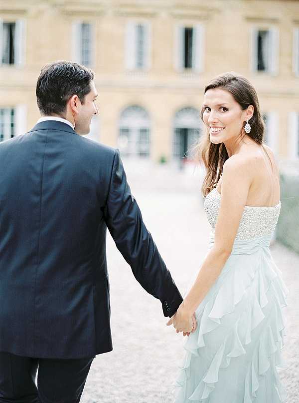A couple portrait shot outdoors on a gravel courtyard in front of a large classical French stone building with tall windows. The groom, seen from behind, wears a dark navy suit, while the bride faces the camera smiling over her shoulder, wearing a strapless gown with a beaded/sequined bodice and a pale ice-blue layered chiffon ruffled skirt. She accessorizes with crystal drop earrings and has her dark hair loosely styled down. The two are holding hands as they walk, creating a candid, mid-movement portrait composition. Potential venue feature image.