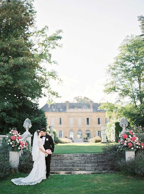 A couple portrait taken outdoors on the formal garden steps of a French chateau, with the honey-colored stone manor house softly out of focus in the background. The bride wears a long-sleeved lace gown with a cathedral-length train and holds a bouquet of coral, hot pink, and white blooms, while the groom is dressed in a classic black tuxedo with a white boutonniere. Two large stone urns flank the steps on either side, each filled with full arrangements of coral dahlias, hot pink blooms, and white flowers creating a symmetrical, classic French formal garden aesthetic. The shot is a wide portrait composition captured in soft, warm natural light with a film-like quality. Potential venue feature image.
