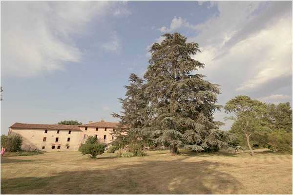 Wide-angle exterior shot of a French country estate or farmhouse-style venue, featuring a large pink-beige rendered stone building visible in the background behind a prominent mature cedar tree. The grounds consist of open lawn with scattered ornamental trees. A figure in what appears to be a pink outfit is barely visible near the building on the far left. The image is taken from a distance, serving primarily as an establishing shot of the property and its grounds. Potential venue feature image.