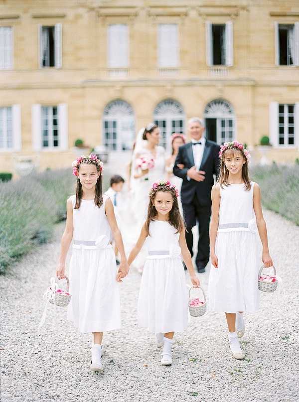 Three flower girls walk along a gravel path toward the camera during an outdoor wedding ceremony procession, with the bride and groom visible in soft focus behind them. The setting is the grounds of a French chateau, with the classical stone facade of the building prominent in the background. The flower girls wear white midi dresses with pale grey sash waistbands, white ankle socks, and white shoes, and each wears a pink floral crown; two carry small wicker baskets filled with pink rose petals. In the background, the bride wears a white ball gown and holds a pink bouquet, while the groom is dressed in a black tuxedo with a bow tie; a young boy and a woman in a pink dress are also visible in the procession. The image is a medium wide shot with a shallow depth of field, keeping the flower girls sharp and the chateau and bridal party softly blurred. Potential venue feature image.