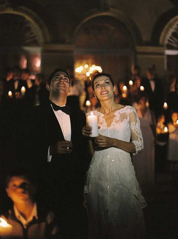 The bride and groom stand together indoors during what appears to be a candlelight ceremony or reception moment, both holding lit white pillar candles and looking upward. The groom wears a black tuxedo with bow tie, while the bride wears a white lace gown with three-quarter sleeves featuring delicate lace overlay and subtle embellishment on the skirt. The room is an ornate interior space with arched architectural detailing and painted ceiling visible in the background, lit by a chandelier and dozens of candles held by guests filling the room behind them. The warm, low candlelight creates deep shadows and golden tones throughout the image, giving it a film photography quality. This is a mid-shot portrait with a shallow depth of field, with guests and their candles softly blurred in the background.