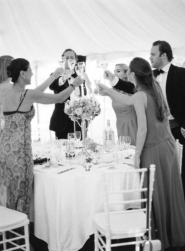 This black-and-white image captures a champagne toast during a wedding reception, with six guests raising their glasses together around a round dining table. The setting is an indoor marquee or tent, identifiable by the draped fabric ceiling visible overhead. The table is dressed with a white linen cloth, crystal glassware, formal place settings, and a tall centerpiece featuring a rounded floral arrangement of roses on what appears to be a candelabra-style stand with hanging pearl or crystal strands. Chiavari chairs surround the table, and a wine cooler is visible in the background. The guests are dressed formally — the men in black tie and the women in floor-length gowns — and the medium-distance portrait shot captures the group mid-toast with high contrast and bright, even tones.