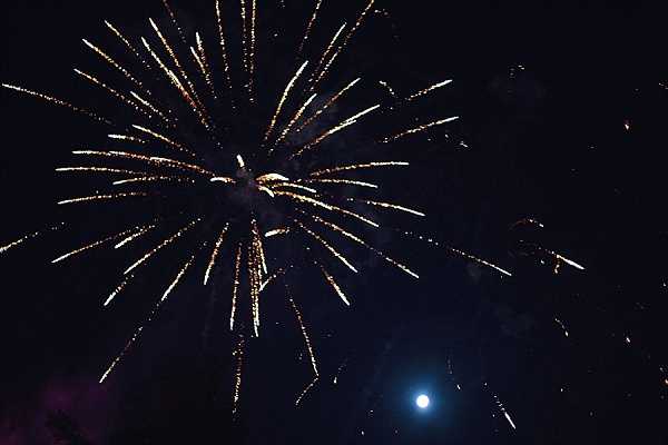 A nighttime fireworks display captured against a dark sky, with a large gold and amber burst dominating the frame and a full moon visible in the lower right corner. A smaller secondary burst is visible to the right, and faint purple/pink smoke or light appears at the lower left edge. No people, venue, or wedding party are visible in this wide shot. The image is slightly underexposed with a dark background, typical of nighttime event photography.