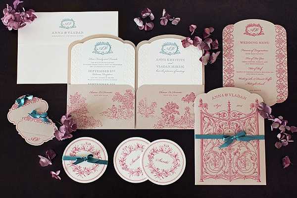 Flat lay photograph of a complete wedding stationery suite spread across a dark background, accented with scattered purple and mauve flower petals. The collection includes a gatefold invitation with trifold panels, an outer pocket envelope in warm ivory/kraft, a wedding menu card with an arched top, circular coasters or favor tags, a place card, and what appears to be a program or itinerary card featuring ornate gate-style die-cut detailing. The design palette is dusty rose/coral pink and teal, with the primary motif being a toile-style illustrated print depicting pastoral French garden scenes, ornate ironwork gates, and floral frames. Teal satin ribbon ties are used on several pieces, and a monogram crest in teal appears on multiple items. The couple's names, Anna Kheyerts and Vladan Mirsic, are visible on the invitation panels. The overall design theme is classic French-inspired with a romantic, vintage aesthetic.