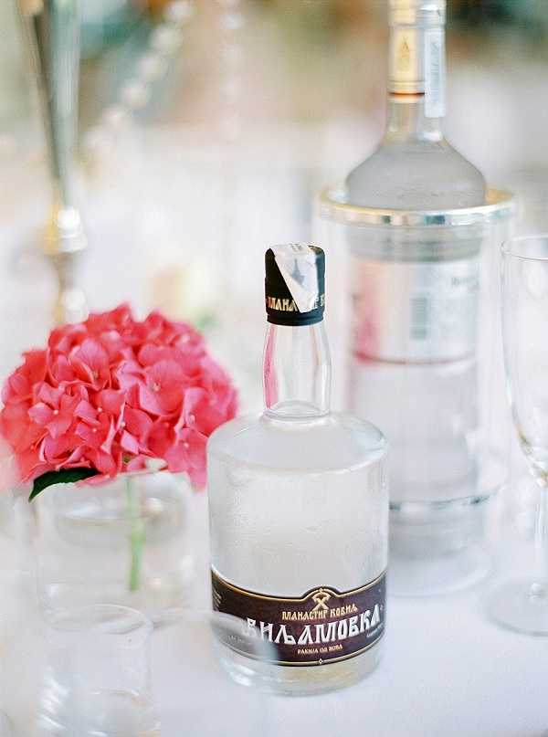 Close-up detail shot of a wedding reception table featuring a bottle of Serbian rakija (Виљамовка/Viljamovka pear brandy) with a dark label and black wax seal in the foreground, alongside a larger clear spirits bottle behind it. A small glass vase holding a single coral-pink hydrangea bloom sits to the left, and clear glassware is partially visible around the bottles. The table linen is white and the overall decor palette is light and airy with pops of coral pink from the floral accent.