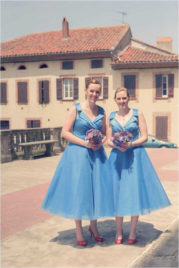Two bridesmaids pose together outdoors on a paved terrace or plaza in front of a traditional French building with terracotta roof tiles and wooden shutters. Both women wear matching cornflower blue tea-length dresses with a 1950s-inspired full skirt silhouette and wide shoulder straps, paired with red peep-toe heels. Each holds a small round bouquet of mixed purple, lavender, and pink flowers including what appear to be asters and cornflowers. The portrait is a medium full-length shot taken in bright midday sunlight.