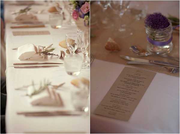 Close-up detail shots of a wedding reception table setting, shown as a side-by-side diptych. The left image shows a long table covered in a white linen tablecloth with place settings featuring kraft paper placecards, white folded napkins garnished with a small sprig of greenery, water glasses, and wooden-handled cutlery. The right image shows a close-up of a kraft paper printed menu card alongside a small glass jar filled with purple allium or amaranth flowers tied with a lavender ribbon, with a seashell visible nearby. The overall decor palette combines ivory, kraft brown, and purple accents, with seashells incorporated as decorative elements suggesting a coastal or beach-inspired styling theme. Both images are detailed/close-up shots with a shallow depth of field.