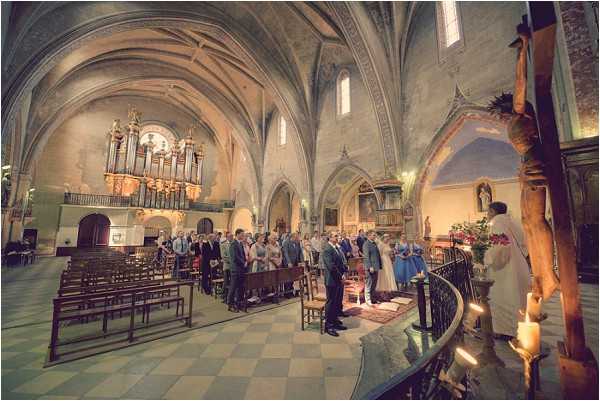 A wedding ceremony taking place inside a historic French Catholic church with vaulted stone ceilings, painted arches, and a large pipe organ visible at the rear of the nave. The bride is wearing a white gown and the groom a dark suit, standing at the altar before a priest in white vestments, with a large wooden crucifix prominent in the foreground right. Bridesmaids in cornflower blue dresses stand alongside the couple, and approximately 40-50 guests are seated and standing in wooden pews. The ceremony is lit by natural light filtering through high windows supplemented by candles near the altar, and a floral arrangement with red and pink flowers decorates the altar rail. Wide-angle shot taken from the side aisle capturing the full depth of the church interior.