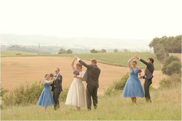 An outdoor bridal party portrait taken in an open countryside setting, with harvested fields and a distant village visible in the background. Three couples are dancing together in a line on a grassy hillside, each pair performing a twirl or spin move. The bride wears a white tea-length dress, while two bridesmaids wear matching cornflower blue tea-length wrap-style dresses with short sleeves, carrying small purple bouquets. The groomsmen and male partners wear dark grey suits. The retro 1950s-inspired styling is reinforced by the tea-length silhouettes and the playful dance poses. Wide shot with a soft, slightly hazy light giving the image a muted, faded tone.