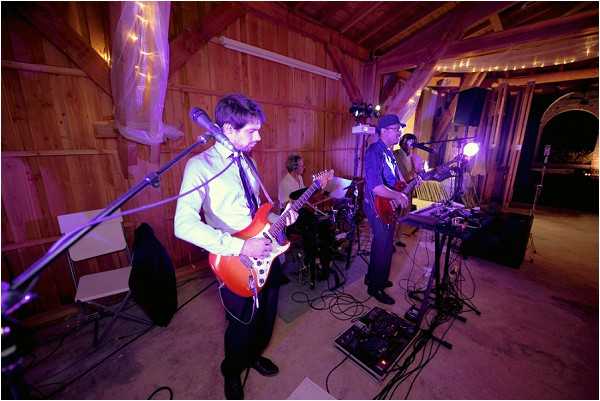 A live band performs at a wedding reception inside a rustic barn venue with natural wood-paneled walls and ceiling. The band consists of at least three visible musicians — a guitarist in the foreground wearing a white dress shirt and dark tie playing a red electric guitar, a second guitarist in a dark suit wearing a cap, and a drummer partially visible in the background. The space is decorated with fairy lights strung along the barn's upper beams and sheer white fabric draped from the rafters, with purple uplighting illuminating the walls. The shot is taken from a wide-angle perspective at floor level, capturing the full performance setup including cables, guitar pedals, a keyboard stand, and audio equipment.
