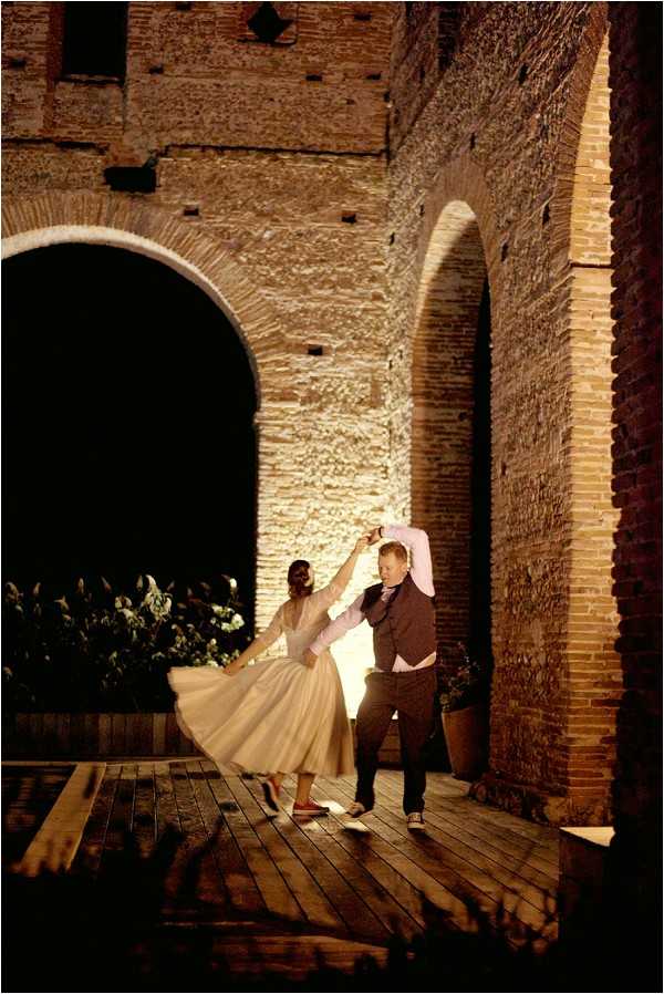A couple dances together at night on a wooden deck beneath large brick archways, likely at a chateau or historic stone building. The bride wears a tea-length ivory dress with long sleeves and an full skirt that is mid-spin, paired with flat shoes, while the groom wears dark trousers, a brown waistcoat, a light shirt, and black sneakers. The scene is dramatically lit by warm uplighting that illuminates the aged brick arches, creating strong contrast against the dark surrounding areas. The portrait-oriented, full-length shot captures a candid, energetic first dance moment with a relaxed, informal styling aesthetic.