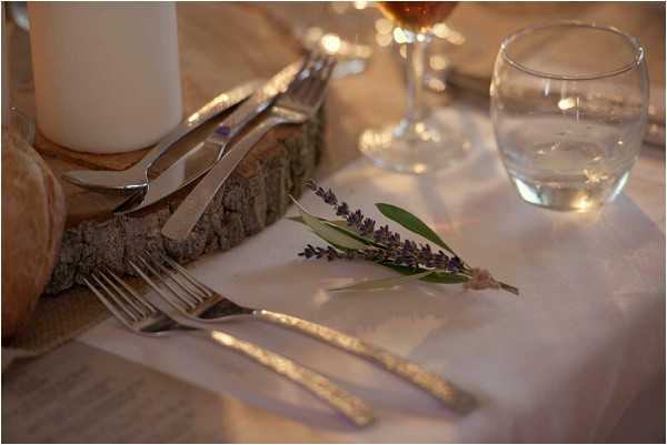 Close-up detail shot of a rustic wedding table setting featuring a small sprig of fresh lavender and sage tied with twine, laid across silver cutlery on a white linen tablecloth. A raw wood slice centerpiece holds a white pillar candle in the background, alongside wine glasses and a water glass. The decor palette combines natural wood, white linen, and purple lavender accents, consistent with a rustic or Provençal styling theme.