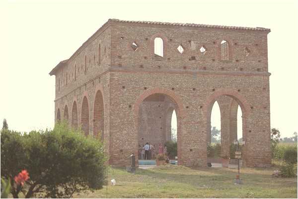 Wide exterior shot of a large two-story open-air brick ruin structure with arched ground-floor openings and decorative diamond and arched window cutouts on the upper level, consistent with a historic or rustic venue setting. A small group of approximately five to eight people, appearing to be wedding guests or bridal party members, are gathered beneath the arches on the ground floor, with what looks like event setup items visible nearby. The image has a soft, slightly washed-out, warm-toned quality suggesting late afternoon or golden hour light. Potential venue feature image.