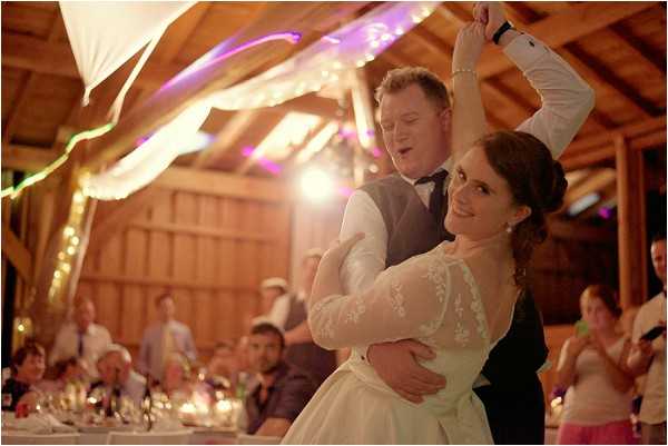 The bride and groom are performing their first dance inside a rustic barn venue, with the groom spinning the bride as she smiles toward the camera. The bride wears a white gown with lace long sleeves, and the groom is dressed in a grey waistcoat, white shirt, and black bow tie. The barn ceiling is decorated with white fabric draping, fairy lights, and purple uplighting along the wooden beams. Seated guests are visible in the background at white-linened reception tables, watching and applauding. The shot is a medium portrait taken at dance floor level, with warm ambient lighting creating a slight lens flare.