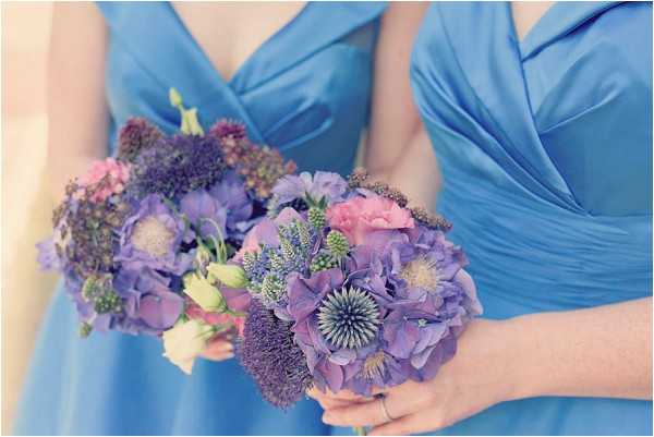 Close-up detail shot of two bridesmaids holding bouquets, with the image cropped to show their torsos and hands. Both bridesmaids are wearing cornflower blue V-neck draped dresses. The bouquets feature purple hydrangeas, blue globe thistles, pink carnations, lavender allium, and small white lisianthus buds, creating a blue-purple and pink color palette. The soft, slightly hazy lighting gives the image a pastel, light-toned quality.