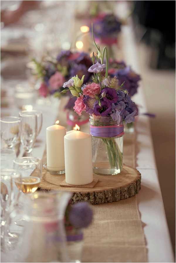Close-up detail shot of a wedding reception table centerpiece with a rustic styling theme. The centerpiece features a mason jar holding a mixed bouquet of purple anemones, lavender hydrangeas, pink carnations, and violet lisianthus, tied with a purple and pink ribbon, arranged on a raw wood slice. Two ivory pillar candles of varying heights sit alongside the floral arrangement on the same wood slice. A neutral burlap table runner runs the length of the long reception table, which is set with wine glasses containing rosé, additional glassware, and repeated floral and candle arrangements visible in the soft-focus background.