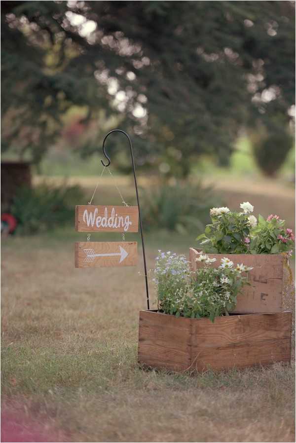 A close-up detail shot of rustic outdoor wedding signage placed on a lawn. Two wooden signs hang from a black shepherd's hook stake — the top sign reads 'Wedding' in white script lettering, and the lower sign displays a white arrow pointing right to direct guests. Beside the stake sits a stacked arrangement of wooden crates used as planters, filled with white, pink, and lavender wildflower-style blooms with green foliage. The overall styling is rustic and bohemian, using natural wood tones and informal flower arrangements as decorative wayfinding elements.