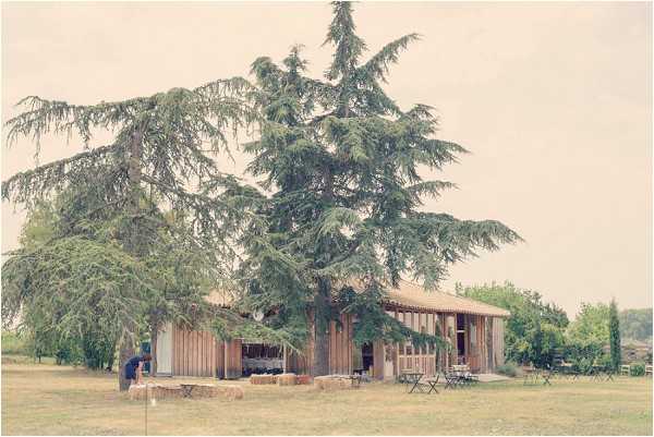 Wide shot of a rustic outdoor wedding venue setup in a rural French setting. The structure is a low, open-sided wooden barn or agricultural building with a thatched or timber roof, partially obscured by a large cedar tree in the foreground. Hay bales are arranged along the front of the building as seating or decorative elements, and bistro-style chairs and small tables are visible to the right side. One or two people can be seen near the entrance, appearing to be setting up. The styling is rustic and informal, consistent with a countryside barn wedding aesthetic. Potential venue feature image.