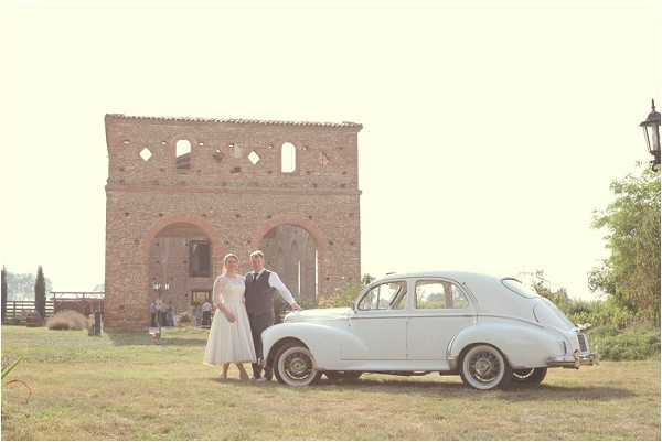 A bride and groom stand together beside a white vintage classic car, positioned outdoors on a grass area in front of a large rustic stone ruin structure with open arched windows and doorways. The bride wears a tea-length white dress with lace sleeves, and the groom is dressed in a dark suit with a waistcoat. The image is taken as a wide portrait shot with warm, hazy golden-hour light giving the scene a slightly overexposed, soft quality. A small group of guests can be seen gathered in the background near the stone structure, suggesting a countryside or rural estate setting. Potential venue feature image.