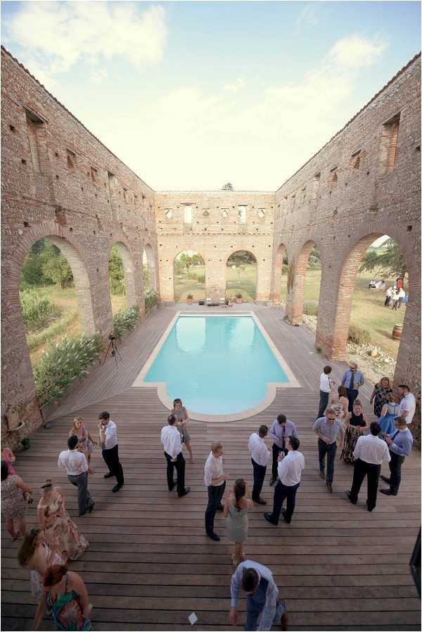 A cocktail hour or reception gathering photographed from an elevated angle, showing approximately 25-30 guests mingling on a wide wooden deck surrounding a rectangular pool. The setting is the roofless interior courtyard of a large ruined stone and brick building with tall arched openings along the walls, open to the countryside beyond. Guests are dressed in smart-casual attire — men in white shirts and dark trousers, women in floral and printed summer dresses. No wedding party or couple is clearly identifiable in the crowd. The decor is minimal, with the architectural ruins and pool serving as the primary visual feature. The wide overhead shot captures the full scale of the courtyard and the social scene below. Potential venue feature image.