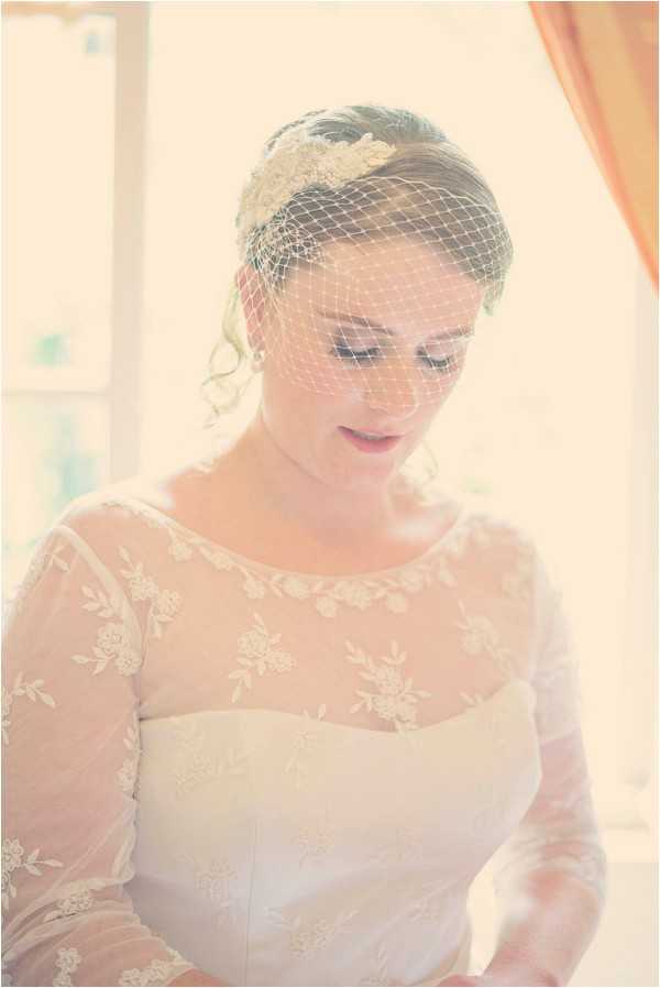 A close-up bridal portrait taken indoors, likely during the getting-ready phase, with soft overexposed natural light from a window in the background giving the image a washed-out, airy, vintage feel. The bride wears an ivory lace wedding dress with long sheer sleeves featuring floral appliqué detailing and a scoop neckline. Her hair is styled in a loose updo and she wears a birdcage veil paired with a pearl and lace hair accessory. The styling has a vintage or retro aesthetic, with the birdcage veil and lace-overlay sleeves as key design elements. The bride is looking downward with a neutral expression.