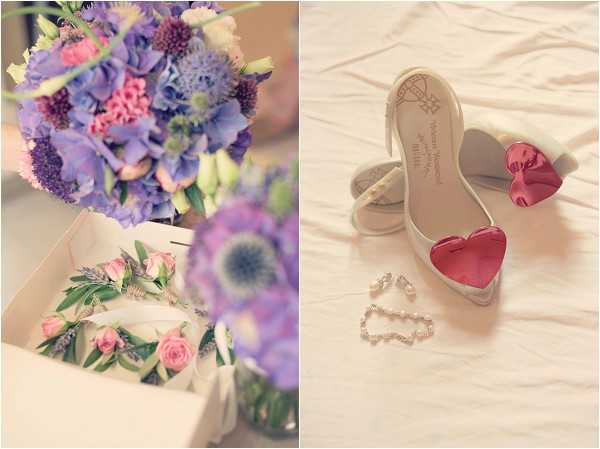 A flat-lay style detail shot composed as a diptych showing bridal accessories. On the left, a bridal bouquet of lavender hydrangeas, purple scabiosa, pink carnations, and blue thistle sits above a white gift box containing a boutonnière with pink rosebuds and greenery, alongside a delicate crystal bracelet. On the right, a pair of white Vivienne Westwood bridal heels with large glossy red heart-shaped embellishments are displayed on white fabric, accompanied by a pearl and crystal drop earring set and a rhinestone bracelet. The overall color palette is soft lavender, pink, and white with pops of red. The image has a warm, slightly hazy pastel tone throughout.