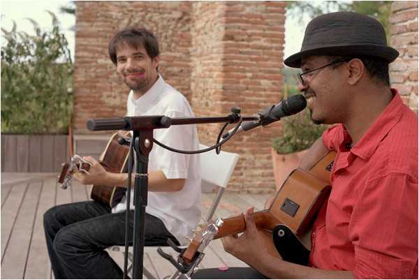 Two male musicians are performing live at what appears to be an outdoor cocktail hour or reception, seated on a wooden deck terrace with an exposed brick wall visible in the background. Both men are playing acoustic guitars; the musician in the foreground wears a red shirt and a dark grey fedora hat and is singing into a microphone, while the second musician in the background wears a white shirt and smiles. The setting appears to be an outdoor terrace of a venue with warm, natural daytime lighting. The shot is a medium close-up taken from a slightly low angle, capturing both performers in the same frame.
