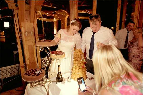 The bride and groom stand together at a dessert table during what appears to be an indoor evening reception, each holding a champagne flute. The bride wears a white lace long-sleeve dress with a hair accessory, and the groom is dressed in a white shirt with a navy blue tie. On the table is a croquembouche — a tower of cream puffs — alongside a separate round cake on a stand and a bottle of champagne. A female guest in the foreground photographs the moment on a smartphone, and two additional guests are visible in the background. The venue features exposed wooden beams and warm string lights overhead, giving the space a rustic barn-style atmosphere. The shot is taken from a slightly elevated angle, capturing the full scene in a wide candid style.