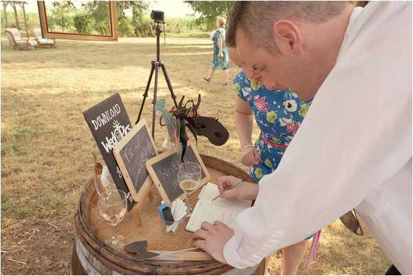 A male guest in a white shirt leans over a rustic wooden barrel used as a cocktail-hour guest book station, writing a message in an open notebook. The barrel is styled with small chalkboard signs on easels — one reads 'Download WeddingPics' — along with two wine glasses and a bottle, giving the setup a casual, rustic feel. A camera on a tripod is positioned nearby, likely set up as a photo booth, and a woman in a blue floral dress walks in the background through an outdoor garden or grounds setting. The composition is a medium close-up shot taken in natural daylight.