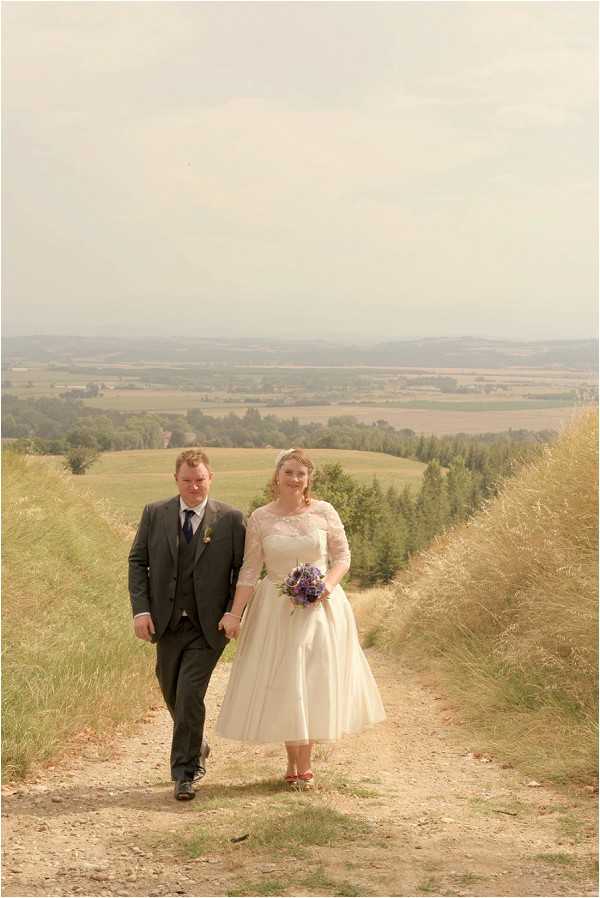 A couple portrait taken outdoors on a hilltop dirt path with a wide panoramic view of rolling agricultural fields and tree lines stretching into the distance behind them. The bride wears a vintage-style tea-length ivory dress with three-quarter lace sleeves and a full skirt, paired with red shoes, and carries a compact round bouquet of purple and blue flowers. The groom wears a dark charcoal three-piece suit with a navy tie and a small yellow boutonniere. The image has a warm, slightly faded tonal quality consistent with vintage-style editing, and is shot as a full-length portrait with the couple walking toward the camera hand in hand.