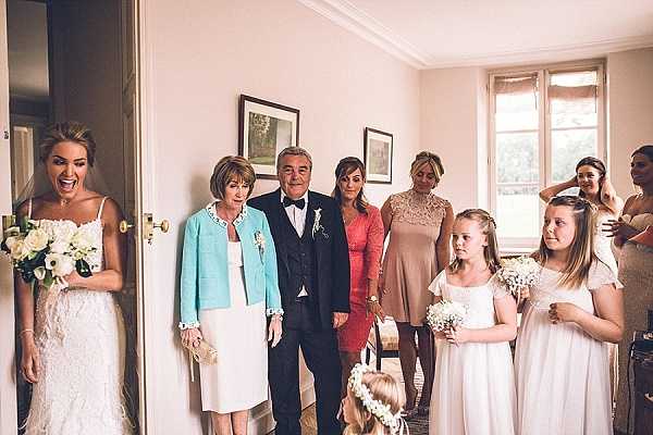 A getting-ready or pre-ceremony moment captured indoors in what appears to be a chateau or manor house room with white walls, crown molding, and tall windows. The bride, wearing a fitted lace gown with thin straps and holding a bouquet of white roses and greenery, is visible in a doorway on the left with an expressive, joyful reaction. Facing her is a group of approximately seven people including a man in a dark suit with bow tie, a woman in a light teal jacket over a white top, a woman in a coral fitted dress, a woman in a taupe lace-overlay dress, two flower girls in white dresses holding small white floral pomanders, and a young girl wearing a floral crown. Additional bridesmaids in blush/nude gowns are partially visible on the far right. The shot is a medium wide-angle composition capturing the candid emotional reveal moment between the bride and her family and bridal party.