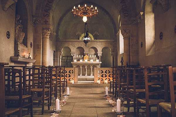 Interior wide shot of a small Romanesque stone chapel set up for a wedding ceremony, with no guests or couple present. The aisle is lined with tall white pillar candles in glass hurricane holders and scattered flower petals, leading toward an ornate stone altar decorated with white candles and what appears to be a gold religious centerpiece. Rows of dark wooden ladder-back chairs flank both sides of the aisle. The altar area is further decorated with clusters of candles along the floor, creating a warm, candlelit atmosphere. An antique wrought-iron chandelier with candle-style bulbs hangs from the arched ceiling, adding to the warm amber lighting throughout the space. The decor palette is white and warm gold, with a classic religious ceremony styling. Potential venue feature image.