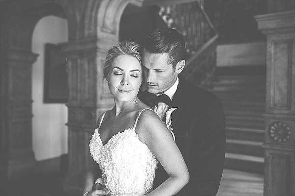 Black-and-white close-up portrait of a bride and groom in an intimate pose inside what appears to be a chateau or manor house, with a grand wooden staircase and ornate carved paneling visible in the softly blurred background. The bride wears a spaghetti-strap gown with textured floral or ruched bodice detailing, her eyes closed as the groom leans in toward her from behind. The groom is dressed in a dark tuxedo with a bow tie. The image has soft contrast with bright highlights on the couple's faces and darker, vignette-style edges, giving it a moody, editorial feel. Portrait composition, mid-shot.