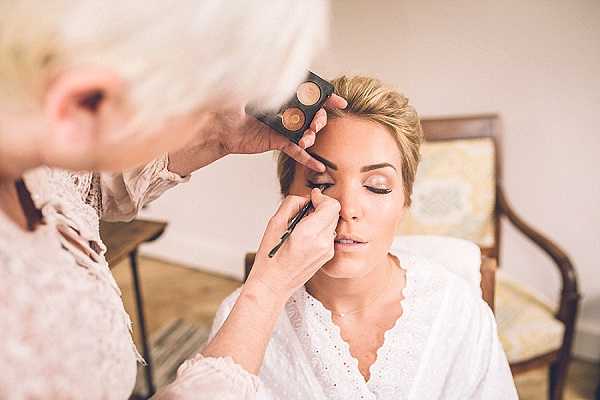 A bride is having her makeup applied during the getting-ready portion of the wedding day. A makeup artist with short white hair is applying eyeliner or eye shadow to the bride's eye using a brush, holding a compact palette in her other hand. The bride has her hair styled up, wears a white lace bridal robe with a scalloped neckline, and has her eyes closed. The setting appears to be an indoor room with a wooden chair and neutral-toned furnishings visible in the background. The shot is a close-up portrait taken from a slight angle, with the makeup artist partially out of frame in the foreground.