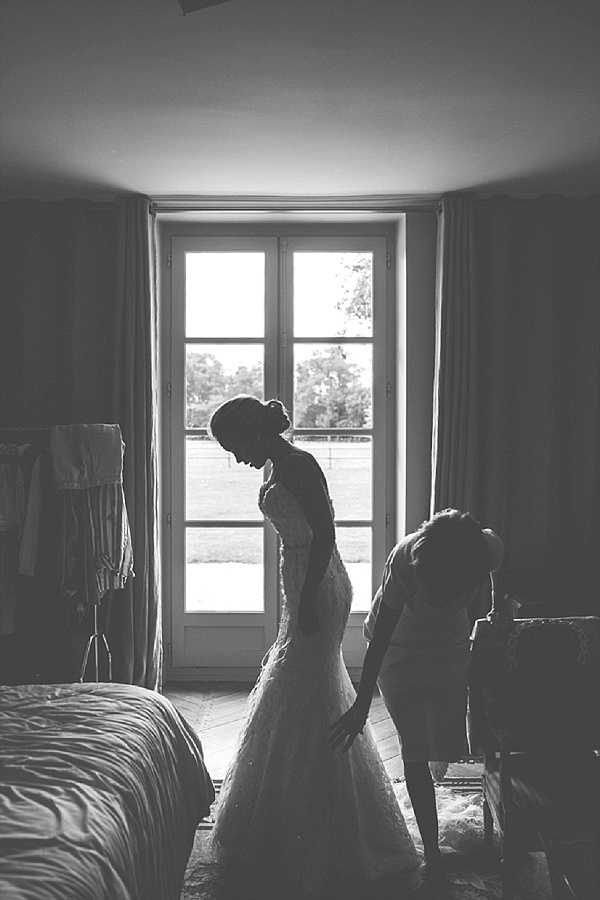 This black-and-white image captures a getting-ready scene indoors in what appears to be a bedroom of a French chateau or manor house, identifiable by the tall French doors, parquet flooring, and draped curtains. The bride, wearing a fitted mermaid-silhouette gown with what appears to be lace or textured fabric detail, stands with her head bowed and hair in an updo, while a second person crouches behind her, likely fastening the dress or attending to the hem. The strong backlight from the French doors creates deep contrast, rendering both figures largely as silhouettes against the bright exterior view. A bed and a garment rack with hanging items are visible on either side of the frame, adding context to the preparation setting. The composition is a medium wide shot with high tonal contrast between the dark interior and the blown-out daylight through the window.