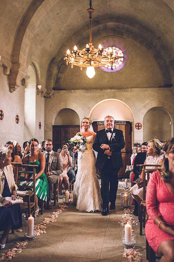A bride is being walked down the aisle by an older man, likely her father, during an indoor church ceremony. The bride wears a fitted, lace mermaid-style gown in ivory and carries a bouquet of white and cream blooms with greenery; the man accompanying her wears a black tuxedo with a white boutonnière. The chapel features vaulted stone arches, a circular stained glass window in red and blue at the rear, and a gold candelabra chandelier overhead, giving the space a Romanesque architectural character. The aisle is lined with tall white pillar candles in glass holders and scattered pale rose petals, while seated guests on wooden chairs on both sides watch the procession — notably a woman in an emerald green dress on the left and a woman in coral pink on the right. The shot is a wide, centered composition taken from the front of the aisle looking back toward the entrance.