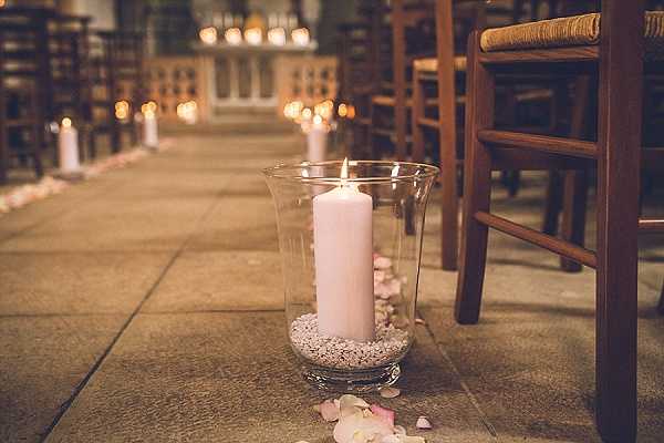 Close-up detail shot of a church ceremony aisle decorated with glass hurricane vases containing pale blush-pink pillar candles set in white pebbles, placed directly on the stone-tiled floor. Scattered pale pink rose petals line the aisle alongside the candles. Wooden rush-seat chairs are visible to the right, and the aisle recedes into the background where rows of matching hurricane candle arrangements and additional candlelight create a warm, glowing ambiance throughout the chapel interior. The overall decor palette is blush pink and warm amber, with a classic, candle-lit ceremony styling.