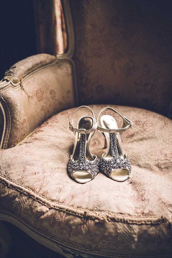 A detail close-up shot of a pair of bridal heels placed on the seat of an ornate vintage armchair with blush pink floral damask upholstery. The shoes are open-toe T-strap sandals with heavily jeweled and beaded embellishment in silver and gunmetal tones across the toe and strap, with a gold interior sole and ankle buckle strap. The warm, moody lighting and vintage chair styling suggest a classic or Old World aesthetic consistent with a French chateau getting-ready setting.