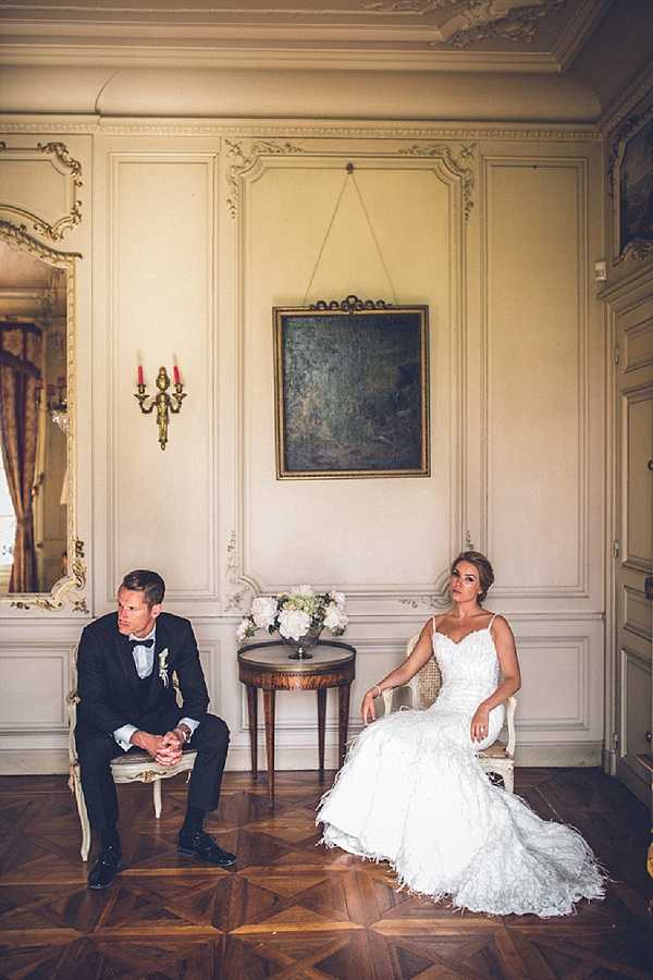 A bride and groom are seated separately on small ornate chairs in an interior room of what appears to be a French chateau, posed apart with a small antique side table between them holding a white floral arrangement of hydrangeas and roses. The groom wears a black tuxedo with a white bow tie and a boutonnière, while the bride wears a fitted, spaghetti-strap lace mermaid gown with a feathered hem. The room features cream boiserie wall paneling with gilded rococo molding details, a gold-framed mirror, a wall-mounted candelabra with red candles, an oil painting hung on the wall, and a parquet wood floor. The portrait is a medium-wide shot with a classic, formal styling aesthetic. Potential venue feature image.