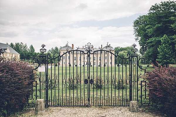 Wide exterior shot of a French chateau viewed through ornate black wrought-iron gates with decorative scrollwork and fleur-de-lis style finials. The gates are set between stone pillars and open onto a gravel path leading across a large lawn to a symmetrical, multi-storey chateau with pale stone facades and tall windows. No people are visible in the frame. The image has a soft, slightly muted color tone with overcast natural light. Potential venue feature image.