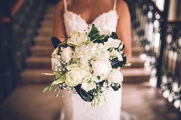 Close-up portrait of a bride holding a rounded bridal bouquet composed of white peonies, white roses, white hydrangea, baby's breath, and dark green foliage. The bride is wearing a white lace spaghetti-strap fitted gown with a sweetheart neckline, visible from the torso up. The background shows an indoor staircase with an ornate dark wood banister, suggesting a chateau or manor house interior. The bouquet is in sharp focus while the bride and background are softly blurred, drawing full attention to the all-white and green floral arrangement.