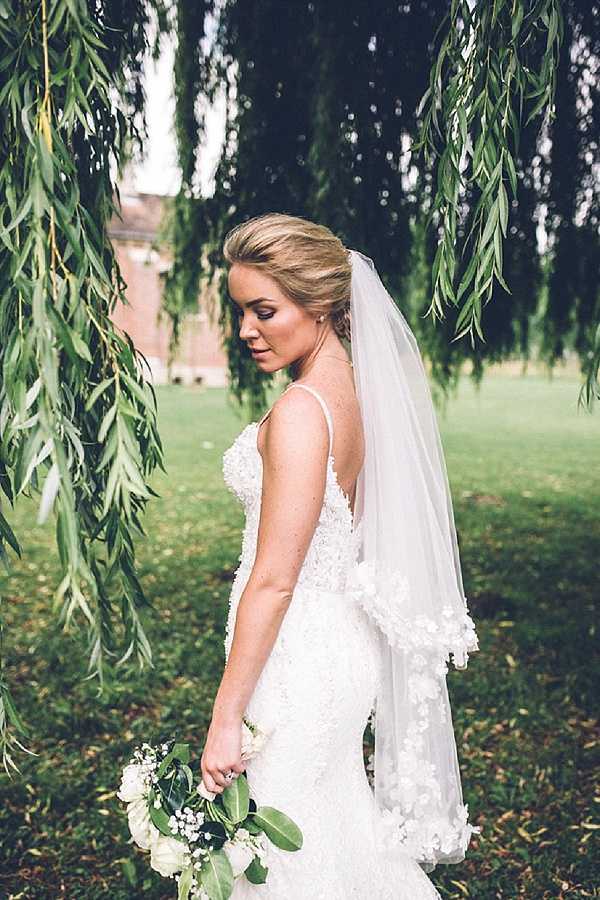 A bridal portrait taken outdoors beneath the hanging branches of a weeping willow tree. The bride wears a fitted white lace gown with spaghetti straps and floral three-dimensional lace appliqué detailing, paired with a mid-length veil trimmed with lace edging. Her blonde hair is styled in a low updo and she looks downward with a relaxed expression. She holds a loosely arranged bouquet of white garden roses, baby's breath, and large green leaves. The composition is a three-quarter portrait shot with the willow branches framing the scene and a red brick building partially visible in the background.