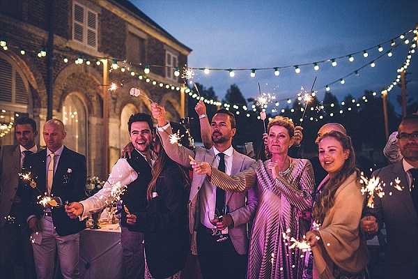 A group of approximately eight wedding guests celebrate outdoors at dusk, holding lit sparklers and laughing during what appears to be a reception moment. The setting is an outdoor courtyard or terrace in front of a brick building, decorated with overhead festoon bulb lights strung across the space. Guests are dressed in cocktail and formal attire including a pink-gold striped dress, a blush suit, dark suits, and a woman wearing a taupe wrap; several guests are holding champagne glasses. The image is a mid-range group shot with warm sparkler light and the deep blue dusk sky creating a contrast with the warm yellow glow of the festoon lights.