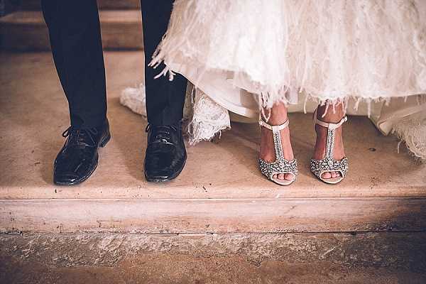 Close-up detail shot of a couple's feet and lower legs standing on stone steps, showing the bride's crystal-embellished ivory T-strap heeled sandals alongside the groom's polished black Oxford lace-up dress shoes. The bottom hem of the bride's white dress is visible, featuring layered tulle with feather trim detailing. The composition is a cropped low-angle detail shot focusing entirely on the footwear and dress hem as a styling detail.