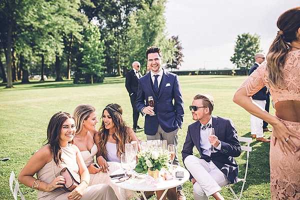 Outdoor cocktail hour scene with a group of approximately six guests gathered around a small white round table on a manicured lawn, with additional guests visible in the background. Three women are seated at the table wearing a blush one-shoulder dress, a strapless champagne dress, and a light lavender dress, while two men stand and sit nearby in navy suits with white trousers. Guests are holding champagne flutes, and the table features a small low floral centerpiece with white and soft green blooms. The setting appears to be a formal garden or chateau grounds, and the overall styling is classic and polished with a neutral blush and navy color palette. Wide shot with a slightly editorial, candid composition.