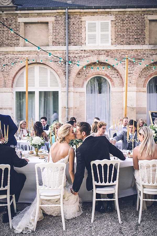 The bride and groom share a kiss while seated at the head of a long outdoor reception dinner table, surrounded by approximately 20-25 guests. The setting is a courtyard in front of a classic French brick building with arched windows and shutters, with teal-colored string lights strung overhead between gold poles. The bride wears a backless lace wedding dress with feather hem details, and the groom is in a black suit. The tablescape features white linen, gold candelabras with tall taper candles, white floral centerpieces including what appear to be white roses or hydrangeas, and gold accents. Guests are seated in white Chiavari-style chairs. The overall decor palette is white, gold, and teal, with a classic French chateau aesthetic. Wide shot taken from slightly behind the couple, capturing both the intimate moment and the full reception scene.