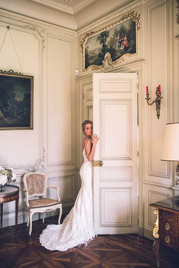 A bridal portrait taken indoors in a French chateau-style room featuring cream-painted boiserie paneling with gold detailing, ornate oil paintings above the doorframes, a gilt wall sconce with red candles, a Louis XV-style caned chair, and a marquetry commode. The bride stands in the doorway, glancing back over her shoulder toward the camera, wearing a fitted ivory lace gown with a low open back, spaghetti straps, and a floor-length train with feathered hem detail. A small white floral arrangement is visible on a side table to the left. The shot is a full-length portrait with a warm, slightly muted color palette, captured from a slight distance to include the full gown and the architectural details of the room. Potential venue feature image.