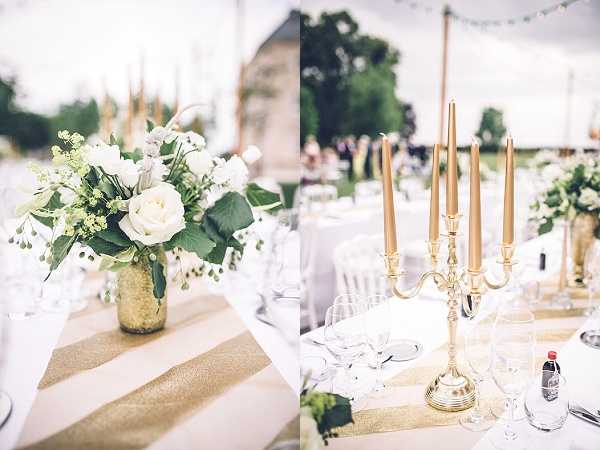 A split close-up detail shot of an outdoor wedding reception table setting with a white and gold color palette. The left panel shows a low centerpiece of white roses, white ranunculus, and green foliage in a gold mercury glass vase, placed on a gold and ivory striped satin table runner over white linen. The right panel features a five-arm gold candelabra with tall gold taper candles, also set on a matching gold striped runner, with crystal wine glasses visible alongside. In the soft-focus background, guests are gathered on a lawn beneath string lights and a white tent structure, suggesting a garden or chateau grounds setting.