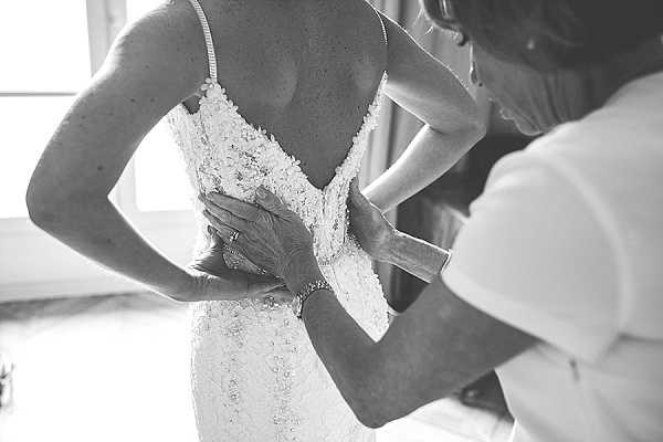 A black-and-white close-up portrait of a bride being helped into her wedding dress by another person, likely a mother or family member. The image is shot indoors with soft natural light coming from a window in the background. The bride's dress features thin spaghetti straps and intricate lace appliqué detailing across the back, with a deep V-back silhouette. The assistant's hands, adorned with a bracelet and ring, are visible fastening or adjusting the back of the gown. The composition is a tight detail shot focused on the backs and hands, emphasizing the texture of the lace against high-contrast monochrome tones.