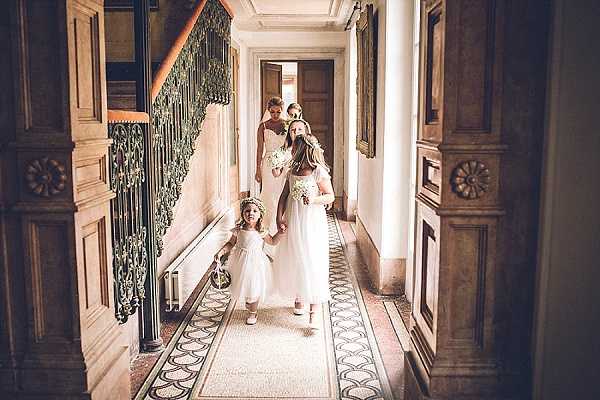 A bridal party walks down a grand interior corridor of what appears to be a chateau or historic mansion, captured in a wide shot from a slightly elevated angle. The procession includes two flower girls in white tea-length dresses with floral crowns, a bridesmaid or junior attendant in a white gown holding a small bouquet of white flowers, and a bridesmaid in a strapless ivory gown visible in the background. The hallway features ornate wood-paneled doors with decorative carved rosettes, a wrought-iron staircase balustrade, and a patterned tile floor runner, all conveying a classic, formal French interior setting. The overall styling is classic and white-toned, with the bridal party dressed uniformly in white and ivory. Potential venue feature image.